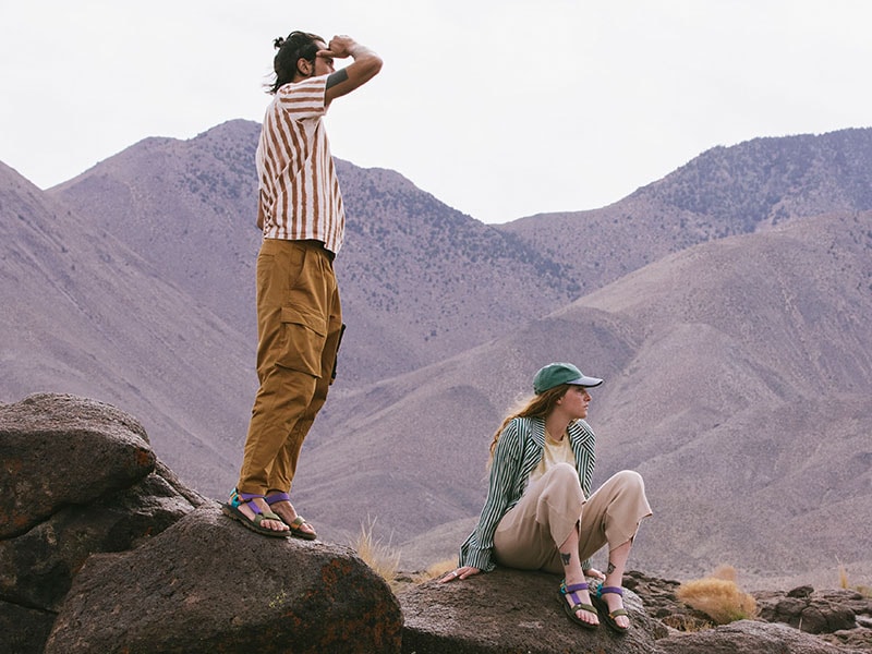 Couple, both wearing Tevas, sitting on a mountaintop and looking out.
