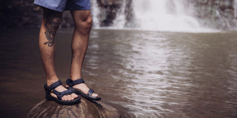 Close up of a person's legs, wearing Teva sandals, standing on a rock in a river.