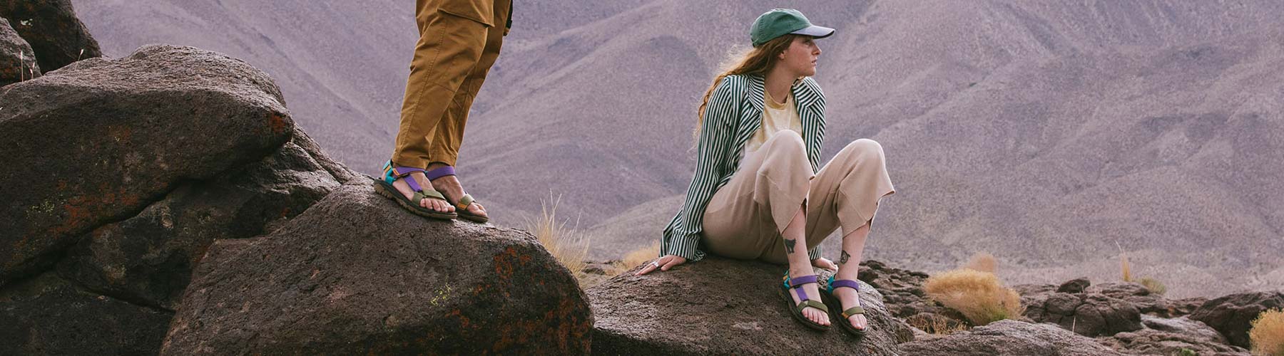 Couple, both wearing Tevas, sitting on a mountaintop and looking out.