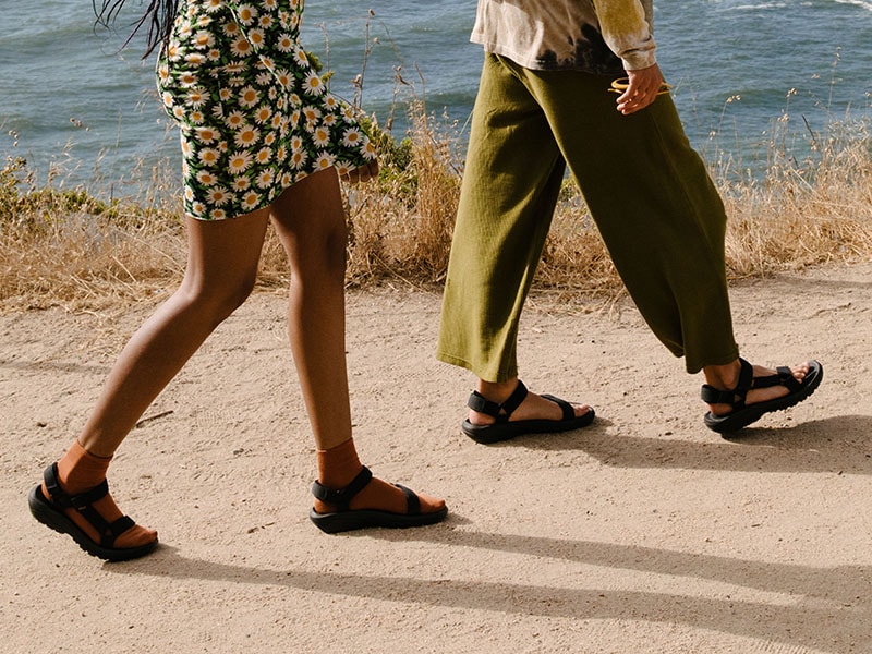 Close up of a man and woman's feet, both wearing Teva sandals on a sand.