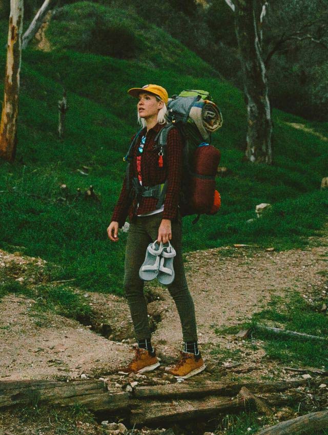 Kate standing in a green field with her hiking pack on, and holding some Tevas.