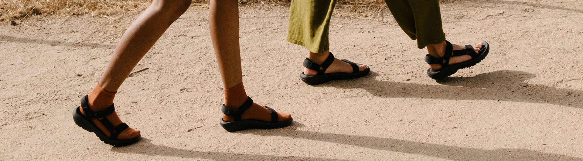 Close up of a man and woman's feet, both wearing Teva sandals on a sand.
