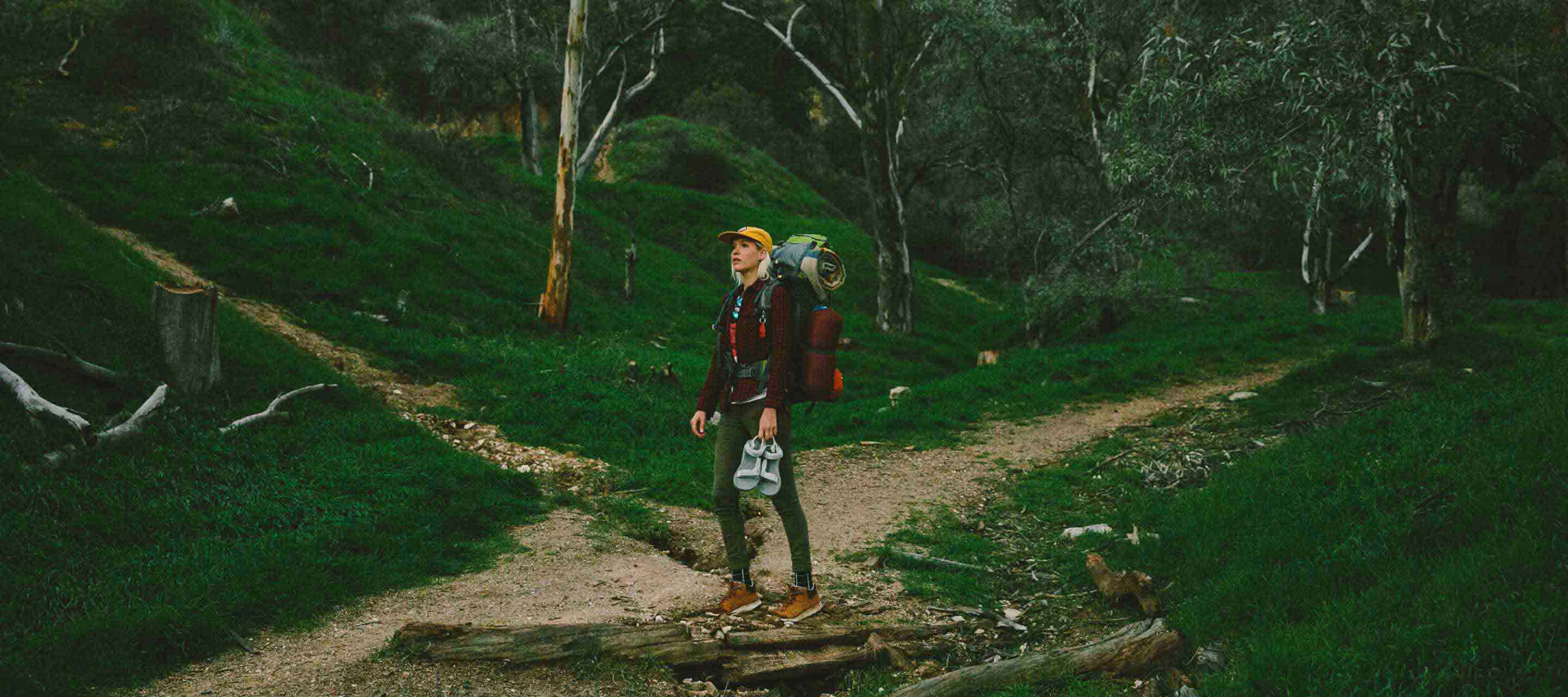 Kate standing in a green field with her hiking pack on, and holding some Tevas.