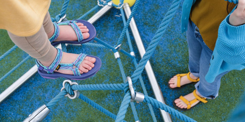 Close up of adult and childs feet, wearing Teva Sandals, standing on a playground.