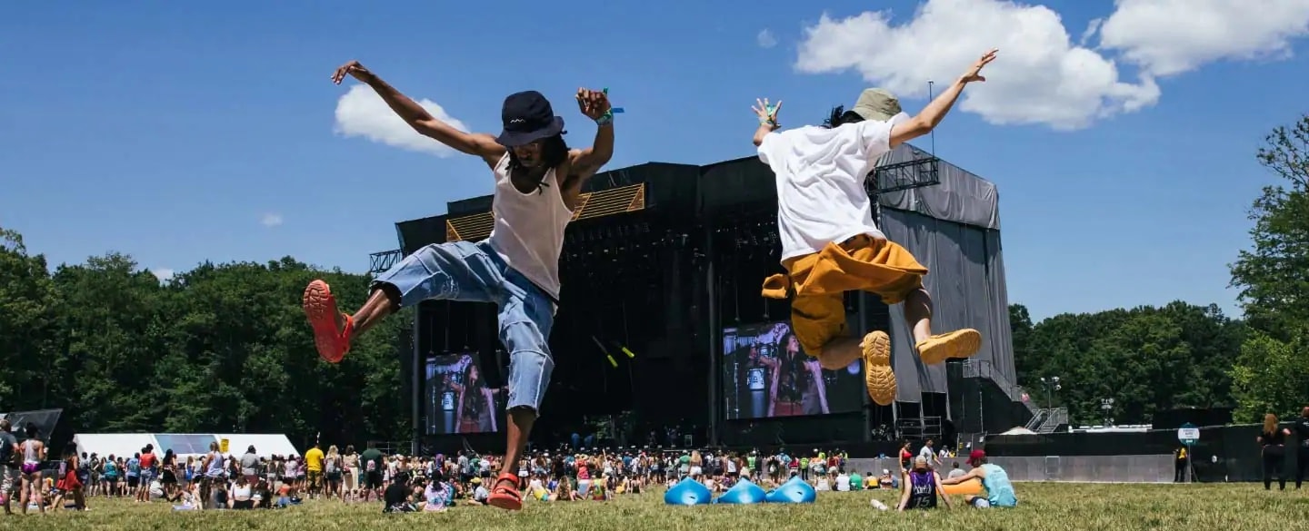 Two guys jumping on lawn in their Tevas, with a stage in the background.